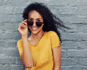 Femme souriante en t-shirt jaune et lunettes de soleil devant un mur gris, mode été Réunion