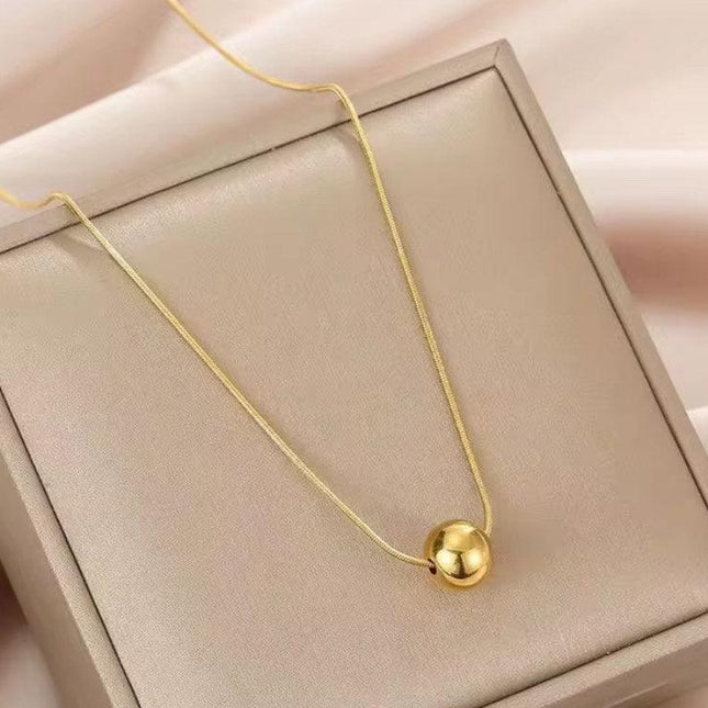 Collier femme doré avec pendentif boule, exposé sur un coffret beige élégant