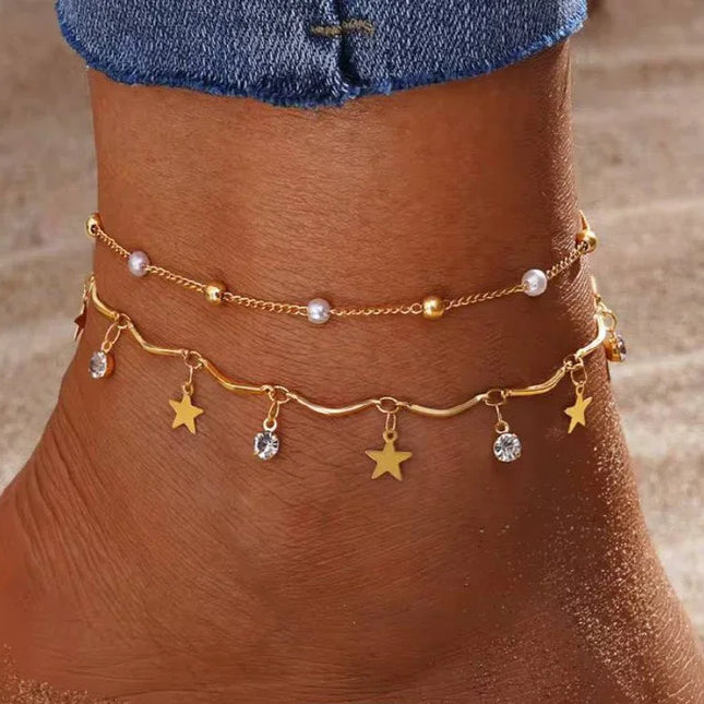 Cheville ornée de deux bracelets dorés, l’un avec perles et l’autre avec pendentifs étoiles et strass, sur fond de sable