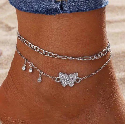 Chaîne de cheville argentée avec pendentif papillon et strass portée sur une cheville bronzée